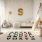 Kinderteppich "Verkehrsalphabet" von spielzwerg. Ambientebild von einem hellen Kinderzimmer mit weißem Holzboden und weißen Wänden, dekoriert mit goldenen Dreiecken und einem großen goldenen Buchstaben 'S'. Links steht ein gemütlicher Sessel mit Kissen und Decke, daneben eine goldene Stehlampe. In der Mitte liegt der Teppich. Rechts sitzt ein lachender Junge in einem weißen Spielzelt mit goldenen Punkten. Im Hintergrund ein schlichtes Bett mit weißer Decke und goldenen Akzenten. RUG-00114-XL, RUG-00114-L.