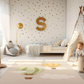 Kinderteppich "Heißluftballons" von spielzwerg. Ambientebild von einem hellen Kinderzimmer mit weißem Holzboden und weißen Wänden, dekoriert mit goldenen Dreiecken und einem großen goldenen Buchstaben 'S'. Links steht ein gemütlicher Sessel mit Kissen und Decke, daneben eine goldene Stehlampe. In der Mitte liegt der Teppich. Rechts sitzt ein lachender Junge in einem weißen Spielzelt mit goldenen Punkten. Im Hintergrund ein schlichtes Bett mit weißer Decke und goldenen Akzenten. RUG-00166-XL, RUG-00166-L.