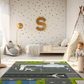 Spielteppich "Flughafen" von spielzwerg. Ambientebild von einem hellen Kinderzimmer mit weißem Holzboden und weißen Wänden, dekoriert mit goldenen Dreiecken und einem großen goldenen Buchstaben 'S'. Links steht ein gemütlicher Sessel mit Kissen und Decke, daneben eine goldene Stehlampe. In der Mitte liegt der Teppich. Rechts sitzt ein lachender Junge in einem weißen Spielzelt mit goldenen Punkten. Im Hintergrund ein schlichtes Bett mit weißer Decke und goldenen Akzenten. RUG-00049-XL, RUG-00049-L.
