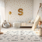 Kinderteppich "Flugverkehr" von spielzwerg. Ambientebild von einem hellen Kinderzimmer mit weißem Holzboden und weißen Wänden, dekoriert mit goldenen Dreiecken und einem großen goldenen Buchstaben 'S'. Links steht ein gemütlicher Sessel mit Kissen und Decke, daneben eine goldene Stehlampe. In der Mitte liegt der Teppich. Rechts sitzt ein lachender Junge in einem weißen Spielzelt mit goldenen Punkten. Im Hintergrund ein schlichtes Bett mit weißer Decke und goldenen Akzenten. RUG-00070-XL, RUG-00070-L.