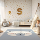 Kinderteppich "Knuffiger Eisbär" von spielzwerg. Ambientebild von einem hellen Kinderzimmer mit weißem Holzboden und weißen Wänden, dekoriert mit goldenen Dreiecken und einem großen goldenen Buchstaben 'S'. Links steht ein gemütlicher Sessel mit Kissen und Decke, daneben eine goldene Stehlampe. In der Mitte liegt der Teppich. Rechts sitzt ein lachender Junge in einem weißen Spielzelt mit goldenen Punkten. Im Hintergrund ein schlichtes Bett mit weißer Decke und goldenen Akzenten. RUG-00153-XL, RUG-00153-L.