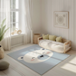 Kinderteppich "Knuffiger Eisbär" von spielzwerg. Ambientebild von einem hellen, freundlichen Kinderzimmer mit weißen Holzwänden und hellem Holzboden. In der Ecke steht ein niedriges Holzbett mit weißen Decken und hellgrünen Kissen. Davor liegt der Teppich. Links steht ein kleines Holzspielzeugrad am Fenster mit weißen Vorhängen. An der Wand hängt ein gerahmtes Bild mit einem dezenten, kreisförmigen Muster. RUG-00153-XL, RUG-00153-L.