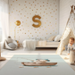 Kinderteppich "Angler-Bär" von spielzwerg. Ambientebild von einem hellen Kinderzimmer mit weißem Holzboden und weißen Wänden, dekoriert mit goldenen Dreiecken und einem großen goldenen Buchstaben 'S'. Links steht ein gemütlicher Sessel mit Kissen und Decke, daneben eine goldene Stehlampe. In der Mitte liegt der Teppich. Rechts sitzt ein lachender Junge in einem weißen Spielzelt mit goldenen Punkten. Im Hintergrund ein schlichtes Bett mit weißer Decke und goldenen Akzenten. RUG-00034-XL, RUG-00034-L.