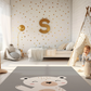 Kinderteppich "Eli Eisbär" von spielzwerg. Ambientebild von einem hellen Kinderzimmer mit weißem Holzboden und weißen Wänden, dekoriert mit goldenen Dreiecken und einem großen goldenen Buchstaben 'S'. Links steht ein gemütlicher Sessel mit Kissen und Decke, daneben eine goldene Stehlampe. In der Mitte liegt der Teppich. Rechts sitzt ein lachender Junge in einem weißen Spielzelt mit goldenen Punkten. Im Hintergrund ein schlichtes Bett mit weißer Decke und goldenen Akzenten. RUG-00123-XL, RUG-00123-L.