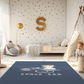 Kinderteppich "Space Bär" von spielzwerg. Ambientebild von einem hellen Kinderzimmer mit weißem Holzboden und weißen Wänden, dekoriert mit goldenen Dreiecken und einem großen goldenen Buchstaben 'S'. Links steht ein gemütlicher Sessel mit Kissen und Decke, daneben eine goldene Stehlampe. In der Mitte liegt der Teppich. Rechts sitzt ein lachender Junge in einem weißen Spielzelt mit goldenen Punkten. Im Hintergrund ein schlichtes Bett mit weißer Decke und goldenen Akzenten. RUG-00048-XL, RUG-00048-L.