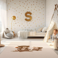 Kinderteppich "Bär und Hörnchen" von spielzwerg. Ambientebild von einem hellen Kinderzimmer mit weißem Holzboden und weißen Wänden, dekoriert mit goldenen Dreiecken und einem großen goldenen Buchstaben 'S'. Links steht ein gemütlicher Sessel mit Kissen und Decke, daneben eine goldene Stehlampe. In der Mitte liegt der Teppich. Rechts sitzt ein lachender Junge in einem weißen Spielzelt mit goldenen Punkten. Im Hintergrund ein schlichtes Bett mit weißer Decke und goldenen Akzenten. RUG-00086-XL, RUG-00086-L.