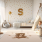 Kinderteppich "Benno Bär" von spielzwerg. Ambientebild von einem hellen Kinderzimmer mit weißem Holzboden und weißen Wänden, dekoriert mit goldenen Dreiecken und einem großen goldenen Buchstaben 'S'. Links steht ein gemütlicher Sessel mit Kissen und Decke, daneben eine goldene Stehlampe. In der Mitte liegt der Teppich. Rechts sitzt ein lachender Junge in einem weißen Spielzelt mit goldenen Punkten. Im Hintergrund ein schlichtes Bett mit weißer Decke und goldenen Akzenten. RUG-00085-XL, RUG-00085-L.