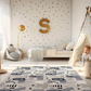 Kinderteppich "Löwenrudel" von spielzwerg. Ambientebild von einem hellen Kinderzimmer mit weißem Holzboden und weißen Wänden, dekoriert mit goldenen Dreiecken und einem großen goldenen Buchstaben 'S'. Links steht ein gemütlicher Sessel mit Kissen und Decke, daneben eine goldene Stehlampe. In der Mitte liegt der Teppich. Rechts sitzt ein lachender Junge in einem weißen Spielzelt mit goldenen Punkten. Im Hintergrund ein schlichtes Bett mit weißer Decke und goldenen Akzenten. RUG-00083-XL, RUG-00083-L.