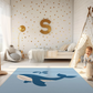 Kinderteppich "Blauwal" von spielzwerg. Ambientebild von einem hellen Kinderzimmer mit weißem Holzboden und weißen Wänden, dekoriert mit goldenen Dreiecken und einem großen goldenen Buchstaben 'S'. Links steht ein gemütlicher Sessel mit Kissen und Decke, daneben eine goldene Stehlampe. In der Mitte liegt der Teppich. Rechts sitzt ein lachender Junge in einem weißen Spielzelt mit goldenen Punkten. Im Hintergrund ein schlichtes Bett mit weißer Decke und goldenen Akzenten. RUG-00206-XL, RUG-00206-L.