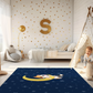 Kinderteppich "Kim Katze" von spielzwerg. Ambientebild von einem hellen Kinderzimmer mit weißem Holzboden und weißen Wänden, dekoriert mit goldenen Dreiecken und einem großen goldenen Buchstaben 'S'. Links steht ein gemütlicher Sessel mit Kissen und Decke, daneben eine goldene Stehlampe. In der Mitte liegt der Teppich. Rechts sitzt ein lachender Junge in einem weißen Spielzelt mit goldenen Punkten. Im Hintergrund ein schlichtes Bett mit weißer Decke und goldenen Akzenten. RUG-00144-XL, RUG-00144-L.
