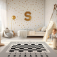 Spielteppich "Schach" von spielzwerg. Ambientebild von einem hellen Kinderzimmer mit weißem Holzboden und weißen Wänden, dekoriert mit goldenen Dreiecken und einem großen goldenen Buchstaben 'S'. Links steht ein gemütlicher Sessel mit Kissen und Decke, daneben eine goldene Stehlampe. In der Mitte liegt der Teppich. Rechts sitzt ein lachender Junge in einem weißen Spielzelt mit goldenen Punkten. Im Hintergrund ein schlichtes Bett mit weißer Decke und goldenen Akzenten. RUG-00010-XL, RUG-00010-L.