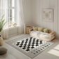 Spielteppich "Schach" von spielzwerg. Ambientebild von einem hellen, freundlichen Kinderzimmer mit weißen Holzwänden und hellem Holzboden. In der Ecke steht ein niedriges Holzbett mit weißen Decken und hellgrünen Kissen. Davor liegt der Teppich. Links steht ein kleines Holzspielzeugrad am Fenster mit weißen Vorhängen. An der Wand hängt ein gerahmtes Bild mit einem dezenten, kreisförmigen Muster. RUG-00010-XL, RUG-00010-L.