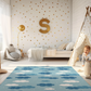 Kinderteppich "Wolkendecke" von spielzwerg. Ambientebild von einem hellen Kinderzimmer mit weißem Holzboden und weißen Wänden, dekoriert mit goldenen Dreiecken und einem großen goldenen Buchstaben 'S'. Links steht ein gemütlicher Sessel mit Kissen und Decke, daneben eine goldene Stehlampe. In der Mitte liegt der Teppich. Rechts sitzt ein lachender Junge in einem weißen Spielzelt mit goldenen Punkten. Im Hintergrund ein schlichtes Bett mit weißer Decke und goldenen Akzenten. RUG-00045-XL, RUG-00045-L.