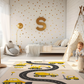 Spielteppich "Baustellenstraße" von spielzwerg. Ambientebild von einem hellen Kinderzimmer mit weißem Holzboden und weißen Wänden, dekoriert mit goldenen Dreiecken und einem großen goldenen Buchstaben 'S'. Links steht ein gemütlicher Sessel mit Kissen und Decke, daneben eine goldene Stehlampe. In der Mitte liegt der Teppich. Rechts sitzt ein lachender Junge in einem weißen Spielzelt mit goldenen Punkten. Im Hintergrund ein schlichtes Bett mit weißer Decke und goldenen Akzenten. RUG-00203-XL, RUG-00203-L.