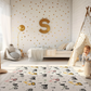 Kinderteppich "Bunte Bagger" von spielzwerg. Ambientebild von einem hellen Kinderzimmer mit weißem Holzboden und weißen Wänden, dekoriert mit goldenen Dreiecken und einem großen goldenen Buchstaben 'S'. Links steht ein gemütlicher Sessel mit Kissen und Decke, daneben eine goldene Stehlampe. In der Mitte liegt der Teppich. Rechts sitzt ein lachender Junge in einem weißen Spielzelt mit goldenen Punkten. Im Hintergrund ein schlichtes Bett mit weißer Decke und goldenen Akzenten. RUG-00125-XL, RUG-00125-L.