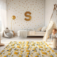 Kinderteppich "Baustellenchaos" von spielzwerg. Ambientebild von einem hellen Kinderzimmer mit weißem Holzboden und weißen Wänden, dekoriert mit goldenen Dreiecken und einem großen goldenen Buchstaben 'S'. Links steht ein gemütlicher Sessel mit Kissen und Decke, daneben eine goldene Stehlampe. In der Mitte liegt der Teppich. Rechts sitzt ein lachender Junge in einem weißen Spielzelt mit goldenen Punkten. Im Hintergrund ein schlichtes Bett mit weißer Decke und goldenen Akzenten. RUG-00069-XL, RUG-00069-L.