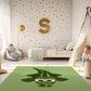 Kinderteppich "Kroko" von spielzwerg. Ambientebild von einem hellen Kinderzimmer mit weißem Holzboden und weißen Wänden, dekoriert mit goldenen Dreiecken und einem großen goldenen Buchstaben 'S'. Links steht ein gemütlicher Sessel mit Kissen und Decke, daneben eine goldene Stehlampe. In der Mitte liegt der Teppich. Rechts sitzt ein lachender Junge in einem weißen Spielzelt mit goldenen Punkten. Im Hintergrund ein schlichtes Bett mit weißer Decke und goldenen Akzenten. RUG-00202-XL, RUG-00202-L.