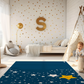 Kinderteppich „Süße Sterne“ von spielzwerg. Ambientebild von einem hellen Kinderzimmer mit weißem Holzboden und weißen Wänden, dekoriert mit goldenen Dreiecken und einem großen goldenen Buchstaben 'S'. Links steht ein gemütlicher Sessel mit Kissen und Decke, daneben eine goldene Stehlampe. In der Mitte liegt der Teppich. Rechts sitzt ein lachender Junge in einem weißen Spielzelt mit goldenen Punkten. Im Hintergrund ein schlichtes Bett mit weißer Decke und goldenen Akzenten. RUG-00158-XL, RUG-00158-L.