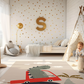 Kinderteppich "Dino im Auto" von spielzwerg. Ambientebild von einem hellen Kinderzimmer mit weißem Holzboden und weißen Wänden, dekoriert mit goldenen Dreiecken und einem großen goldenen Buchstaben 'S'. Links steht ein gemütlicher Sessel mit Kissen und Decke, daneben eine goldene Stehlampe. In der Mitte liegt der Teppich. Rechts sitzt ein lachender Junge in einem weißen Spielzelt mit goldenen Punkten. Im Hintergrund ein schlichtes Bett mit weißer Decke und goldenen Akzenten. RUG-00030-XL, RUG-00030-L.