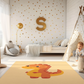 Kinderteppich "Lino Dino" von spielzwerg. Ambientebild von einem hellen Kinderzimmer mit weißem Holzboden und weißen Wänden, dekoriert mit goldenen Dreiecken und einem großen goldenen Buchstaben 'S'. Links steht ein gemütlicher Sessel mit Kissen und Decke, daneben eine goldene Stehlampe. In der Mitte liegt der Teppich. Rechts sitzt ein lachender Junge in einem weißen Spielzelt mit goldenen Punkten. Im Hintergrund ein schlichtes Bett mit weißer Decke und goldenen Akzenten. RUG-00110-XL, RUG-00110-L.