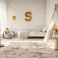 Kinderteppich "Dino-Landschaft" von spielzwerg. Ambientebild von einem hellen Kinderzimmer mit weißem Holzboden und weißen Wänden, dekoriert mit goldenen Dreiecken und einem großen goldenen Buchstaben 'S'. Links steht ein gemütlicher Sessel mit Kissen und Decke, daneben eine goldene Stehlampe. In der Mitte liegt der Teppich. Rechts sitzt ein lachender Junge in einem weißen Spielzelt mit goldenen Punkten. Im Hintergrund ein schlichtes Bett mit weißer Decke und goldenen Akzenten. RUG-00092-XL, RUG-00092-L.