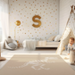 Kinderteppich "Dino-Skelett" von spielzwerg. Ambientebild von einem hellen Kinderzimmer mit weißem Holzboden und weißen Wänden, dekoriert mit goldenen Dreiecken und einem großen goldenen Buchstaben 'S'. Links steht ein gemütlicher Sessel mit Kissen und Decke, daneben eine goldene Stehlampe. In der Mitte liegt der Teppich. Rechts sitzt ein lachender Junge in einem weißen Spielzelt mit goldenen Punkten. Im Hintergrund ein schlichtes Bett mit weißer Decke und goldenen Akzenten. RUG-00174-XL, RUG-00174-L.