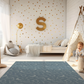 Kinderteppich "Kreidezeit" von spielzwerg. Ambientebild von einem hellen Kinderzimmer mit weißem Holzboden und weißen Wänden, dekoriert mit goldenen Dreiecken und einem großen goldenen Buchstaben 'S'. Links steht ein gemütlicher Sessel mit Kissen und Decke, daneben eine goldene Stehlampe. In der Mitte liegt der Teppich. Rechts sitzt ein lachender Junge in einem weißen Spielzelt mit goldenen Punkten. Im Hintergrund ein schlichtes Bett mit weißer Decke und goldenen Akzenten. RUG-00028-XL, RUG-00028-L.