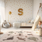 Kinderteppich "Niedliche Dinos" von spielzwerg. Ambientebild von einem hellen Kinderzimmer mit weißem Holzboden und weißen Wänden, dekoriert mit goldenen Dreiecken und einem großen goldenen Buchstaben 'S'. Links steht ein gemütlicher Sessel mit Kissen und Decke, daneben eine goldene Stehlampe. In der Mitte liegt der Teppich. Rechts sitzt ein lachender Junge in einem weißen Spielzelt mit goldenen Punkten. Im Hintergrund ein schlichtes Bett mit weißer Decke und goldenen Akzenten. RUG-00081-XL, RUG-00081-L.
