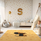 Kinderteppich "Danny Dackel" von spielzwerg. Ambientebild von einem hellen Kinderzimmer mit weißem Holzboden und weißen Wänden, dekoriert mit goldenen Dreiecken und einem großen goldenen Buchstaben 'S'. Links steht ein gemütlicher Sessel mit Kissen und Decke, daneben eine goldene Stehlampe. In der Mitte liegt der Teppich. Rechts sitzt ein lachender Junge in einem weißen Spielzelt mit goldenen Punkten. Im Hintergrund ein schlichtes Bett mit weißer Decke und goldenen Akzenten. RUG-00146-XL, RUG-00146-L.