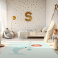 Kinderteppich "Flipper" von spielzwerg. Ambientebild von einem hellen Kinderzimmer mit weißem Holzboden und weißen Wänden, dekoriert mit goldenen Dreiecken und einem großen goldenen Buchstaben 'S'. Links steht ein gemütlicher Sessel mit Kissen und Decke, daneben eine goldene Stehlampe. In der Mitte liegt der Teppich. Rechts sitzt ein lachender Junge in einem weißen Spielzelt mit goldenen Punkten. Im Hintergrund ein schlichtes Bett mit weißer Decke und goldenen Akzenten. RUG-00201-XL, RUG-00201-L.