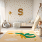 Kinderteppich "Feuerdrache" von spielzwerg. Ambientebild von einem hellen Kinderzimmer mit weißem Holzboden und weißen Wänden, dekoriert mit goldenen Dreiecken und einem großen goldenen Buchstaben 'S'. Links steht ein gemütlicher Sessel mit Kissen und Decke, daneben eine goldene Stehlampe. In der Mitte liegt der Teppich. Rechts sitzt ein lachender Junge in einem weißen Spielzelt mit goldenen Punkten. Im Hintergrund ein schlichtes Bett mit weißer Decke und goldenen Akzenten. RUG-00006-XL, RUG-00006-L.