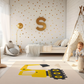 Kinderteppich "Großer Bagger" von spielzwerg. Ambientebild von einem hellen Kinderzimmer mit weißem Holzboden und weißen Wänden, dekoriert mit goldenen Dreiecken und einem großen goldenen Buchstaben 'S'. Links steht ein gemütlicher Sessel mit Kissen und Decke, daneben eine goldene Stehlampe. In der Mitte liegt der Teppich. Rechts sitzt ein lachender Junge in einem weißen Spielzelt mit goldenen Punkten. Im Hintergrund ein schlichtes Bett mit weißer Decke und goldenen Akzenten. RUG-00200-XL, RUG-00200-L.