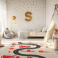Spielteppich "Feuerwehreinsatz" von spielzwerg. Ambientebild von einem hellen Kinderzimmer mit weißem Holzboden und weißen Wänden, dekoriert mit goldenen Dreiecken und einem großen goldenen Buchstaben 'S'. Links steht ein gemütlicher Sessel mit Kissen und Decke, daneben eine goldene Stehlampe. In der Mitte liegt der Teppich. Rechts sitzt ein lachender Junge in einem weißen Spielzelt mit goldenen Punkten. Im Hintergrund ein schlichtes Bett mit weißer Decke und goldenen Akzenten. RUG-00199-XL, RUG-00199-L.