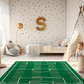Spielteppich "Fußballfeld" von spielzwerg. Ambientebild von einem hellen Kinderzimmer mit weißem Holzboden und weißen Wänden, dekoriert mit goldenen Dreiecken und einem großen goldenen Buchstaben 'S'. Links steht ein gemütlicher Sessel mit Kissen und Decke, daneben eine goldene Stehlampe. In der Mitte liegt der Teppich. Rechts sitzt ein lachender Junge in einem weißen Spielzelt mit goldenen Punkten. Im Hintergrund ein schlichtes Bett mit weißer Decke und goldenen Akzenten. RUG-00121-XL, RUG-00121-L.