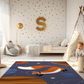 Kinderteppich "Fuchs im All" von spielzwerg. Ambientebild von einem hellen Kinderzimmer mit weißem Holzboden und weißen Wänden, dekoriert mit goldenen Dreiecken und einem großen goldenen Buchstaben 'S'. Links steht ein gemütlicher Sessel mit Kissen und Decke, daneben eine goldene Stehlampe. In der Mitte liegt der Teppich. Rechts sitzt ein lachender Junge in einem weißen Spielzelt mit goldenen Punkten. Im Hintergrund ein schlichtes Bett mit weißer Decke und goldenen Akzenten. RUG-00026-XL, RUG-00026-L.