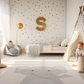 Kinderteppich „Kleiner Elefant“ von spielzwerg. Ambientebild von einem hellen Kinderzimmer mit weißem Holzboden und weißen Wänden, dekoriert mit goldenen Dreiecken und einem großen goldenen Buchstaben 'S'. Links steht ein gemütlicher Sessel mit Kissen und Decke, daneben eine goldene Stehlampe. In der Mitte liegt der Teppich. Rechts sitzt ein lachender Junge in einem weißen Spielzelt mit goldenen Punkten. Im Hintergrund ein schlichtes Bett mit weißer Decke und goldenen Akzenten. RUG-00196-XL, RUG-00196-L.