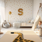 Kinderteppich "Guido Giraffe" von spielzwerg. Ambientebild von einem hellen Kinderzimmer mit weißem Holzboden und weißen Wänden, dekoriert mit goldenen Dreiecken und einem großen goldenen Buchstaben 'S'. Links steht ein gemütlicher Sessel mit Kissen und Decke, daneben eine goldene Stehlampe. In der Mitte liegt der Teppich. Rechts sitzt ein lachender Junge in einem weißen Spielzelt mit goldenen Punkten. Im Hintergrund ein schlichtes Bett mit weißer Decke und goldenen Akzenten. RUG-00057-XL, RUG-00057-L.