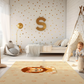 Kinderteppich "Leon Löwe" von spielzwerg. Ambientebild von einem hellen Kinderzimmer mit weißem Holzboden und weißen Wänden, dekoriert mit goldenen Dreiecken und einem großen goldenen Buchstaben 'S'. Links steht ein gemütlicher Sessel mit Kissen und Decke, daneben eine goldene Stehlampe. In der Mitte liegt der Teppich. Rechts sitzt ein lachender Junge in einem weißen Spielzelt mit goldenen Punkten. Im Hintergrund ein schlichtes Bett mit weißer Decke und goldenen Akzenten. RUG-00150-XL, RUG-00150-L.