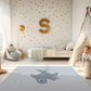 Kinderteppich "Süßer Hai" von spielzwerg. Ambientebild von einem hellen Kinderzimmer mit weißem Holzboden und weißen Wänden, dekoriert mit goldenen Dreiecken und einem großen goldenen Buchstaben 'S'. Links steht ein gemütlicher Sessel mit Kissen und Decke, daneben eine goldene Stehlampe. In der Mitte liegt der Teppich. Rechts sitzt ein lachender Junge in einem weißen Spielzelt mit goldenen Punkten. Im Hintergrund ein schlichtes Bett mit weißer Decke und goldenen Akzenten. RUG-00195-XL, RUG-00195-L.