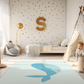 Kinderteppich "Walter" von spielzwerg. Ambientebild von einem hellen Kinderzimmer mit weißem Holzboden und weißen Wänden, dekoriert mit goldenen Dreiecken und einem großen goldenen Buchstaben 'S'. Links steht ein gemütlicher Sessel mit Kissen und Decke, daneben eine goldene Stehlampe. In der Mitte liegt der Teppich. Rechts sitzt ein lachender Junge in einem weißen Spielzelt mit goldenen Punkten. Im Hintergrund ein schlichtes Bett mit weißer Decke und goldenen Akzenten. RUG-00193-XL, RUG-00193-L.