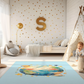 Kinderteppich "Unsere Erde" von spielzwerg. Ambientebild von einem hellen Kinderzimmer mit weißem Holzboden und weißen Wänden, dekoriert mit goldenen Dreiecken und einem großen goldenen Buchstaben 'S'. Links steht ein gemütlicher Sessel mit Kissen und Decke, daneben eine goldene Stehlampe. In der Mitte liegt der Teppich. Rechts sitzt ein lachender Junge in einem weißen Spielzelt mit goldenen Punkten. Im Hintergrund ein schlichtes Bett mit weißer Decke und goldenen Akzenten. RUG-00169-XL, RUG-00169-L.