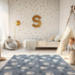 Kinderteppich "Eisbärenlandschaft" von spielzwerg. Ambientebild von einem hellen Kinderzimmer mit weißem Holzboden und weißen Wänden, dekoriert mit goldenen Dreiecken und einem großen goldenen Buchstaben 'S'. Links steht ein gemütlicher Sessel mit Kissen und Decke, daneben eine goldene Stehlampe. In der Mitte liegt der Teppich. Rechts sitzt ein lachender Junge in einem weißen Spielzelt mit goldenen Punkten. Im Hintergrund ein schlichtes Bett mit weißer Decke und goldenen Akzenten. RUG-00140-XL, RUG-00140-L.