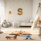 Kinderteppich "Tapferer Ritter" von spielzwerg. Ambientebild von einem hellen Kinderzimmer mit weißem Holzboden und weißen Wänden, dekoriert mit goldenen Dreiecken und einem großen goldenen Buchstaben 'S'. Links steht ein gemütlicher Sessel mit Kissen und Decke, daneben eine goldene Stehlampe. In der Mitte liegt der Teppich. Rechts sitzt ein lachender Junge in einem weißen Spielzelt mit goldenen Punkten. Im Hintergrund ein schlichtes Bett mit weißer Decke und goldenen Akzenten. RUG-00208-XL, RUG-00208-L.