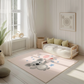 Kinderteppich "Cora Koala" von spielzwerg. Ambientebild von einem hellen, freundlichen Kinderzimmer mit weißen Holzwänden und hellem Holzboden. In der Ecke steht ein niedriges Holzbett mit weißen Decken und hellgrünen Kissen. Davor liegt der Teppich. Links steht ein kleines Holzspielzeugrad am Fenster mit weißen Vorhängen. An der Wand hängt ein gerahmtes Bild mit einem dezenten, kreisförmigen Muster. RUG-00022-XL, RUG-00022-L.