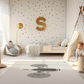 Kinderteppich "Koala-Gesicht" von spielzwerg. Ambientebild von einem hellen Kinderzimmer mit weißem Holzboden und weißen Wänden, dekoriert mit goldenen Dreiecken und einem großen goldenen Buchstaben 'S'. Links steht ein gemütlicher Sessel mit Kissen und Decke, daneben eine goldene Stehlampe. In der Mitte liegt der Teppich. Rechts sitzt ein lachender Junge in einem weißen Spielzelt mit goldenen Punkten. Im Hintergrund ein schlichtes Bett mit weißer Decke und goldenen Akzenten. RUG-00191-XL, RUG-00191-L.