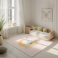 Kinderteppich "Maja Koala" von spielzwerg. Ambientebild von einem hellen, freundlichen Kinderzimmer mit weißen Holzwänden und hellem Holzboden. In der Ecke steht ein niedriges Holzbett mit weißen Decken und hellgrünen Kissen. Davor liegt der Teppich. Links steht ein kleines Holzspielzeugrad am Fenster mit weißen Vorhängen. An der Wand hängt ein gerahmtes Bild mit einem dezenten, kreisförmigen Muster. RUG-00095-XL, RUG-00095-L.
