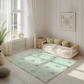 Kinderteppich "Koalas im Eukalyptus" von spielzwerg. Ambientebild von einem hellen, freundlichen Kinderzimmer mit weißen Holzwänden und hellem Holzboden. In der Ecke steht ein niedriges Holzbett mit weißen Decken und hellgrünen Kissen. Davor liegt der Teppich. Links steht ein kleines Holzspielzeugrad am Fenster mit weißen Vorhängen. An der Wand hängt ein gerahmtes Bild mit einem dezenten, kreisförmigen Muster. RUG-00021-XL, RUG-00021-L.