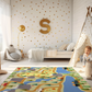 Spielteppich "Gebirgspass" von spielzwerg. Ambientebild von einem hellen Kinderzimmer mit weißem Holzboden und weißen Wänden, dekoriert mit goldenen Dreiecken und einem großen goldenen Buchstaben 'S'. Links steht ein gemütlicher Sessel mit Kissen und Decke, daneben eine goldene Stehlampe. In der Mitte liegt der Teppich. Rechts sitzt ein lachender Junge in einem weißen Spielzelt mit goldenen Punkten. Im Hintergrund ein schlichtes Bett mit weißer Decke und goldenen Akzenten. RUG-00036-XL, RUG-00036-L.