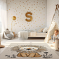 Kinderteppich "Simba" von spielzwerg. Ambientebild von einem hellen Kinderzimmer mit weißem Holzboden und weißen Wänden, dekoriert mit goldenen Dreiecken und einem großen goldenen Buchstaben 'S'. Links steht ein gemütlicher Sessel mit Kissen und Decke, daneben eine goldene Stehlampe. In der Mitte liegt der Teppich. Rechts sitzt ein lachender Junge in einem weißen Spielzelt mit goldenen Punkten. Im Hintergrund ein schlichtes Bett mit weißer Decke und goldenen Akzenten. RUG-00096-XL, RUG-00096-L.