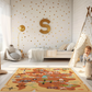 Spielteppich "Schatzjagd" von spielzwerg. Ambientebild von einem hellen Kinderzimmer mit weißem Holzboden und weißen Wänden, dekoriert mit goldenen Dreiecken und einem großen goldenen Buchstaben 'S'. Links steht ein gemütlicher Sessel mit Kissen und Decke, daneben eine goldene Stehlampe. In der Mitte liegt der Teppich. Rechts sitzt ein lachender Junge in einem weißen Spielzelt mit goldenen Punkten. Im Hintergrund ein schlichtes Bett mit weißer Decke und goldenen Akzenten. RUG-00098-XL, RUG-00098-L.